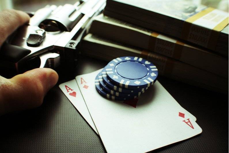 Person holding gun on table with cards, chips, and cash